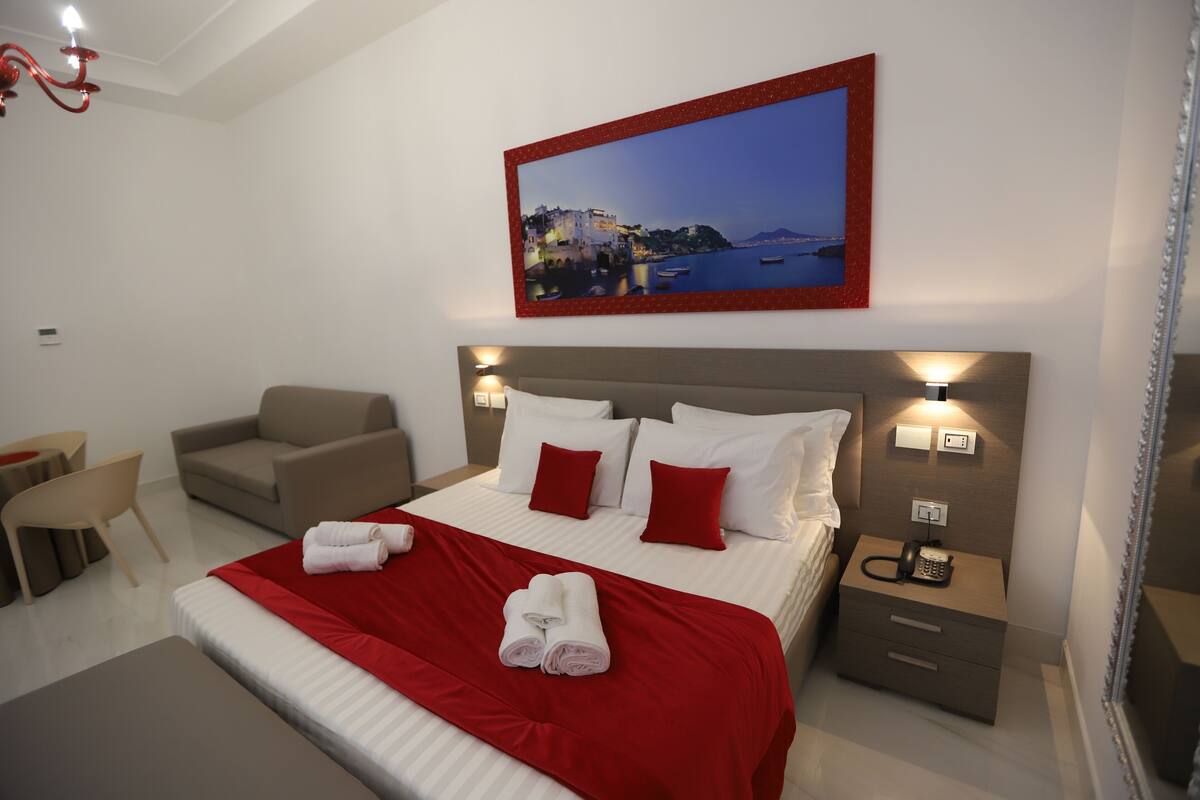Ein modernes Hotelzimmer mit einem großen Bett, roten und weißen Bettwaren, einem Nachttisch mit Telefon, einem Sessel und einem Wandbild mit Küstenlandschaft.