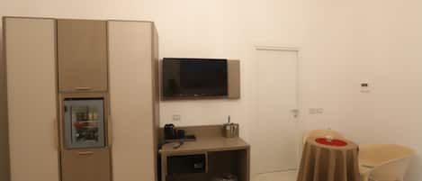 A compact room with a kitchenette, a TV mounted on the wall, a small table with a chair, and a door.