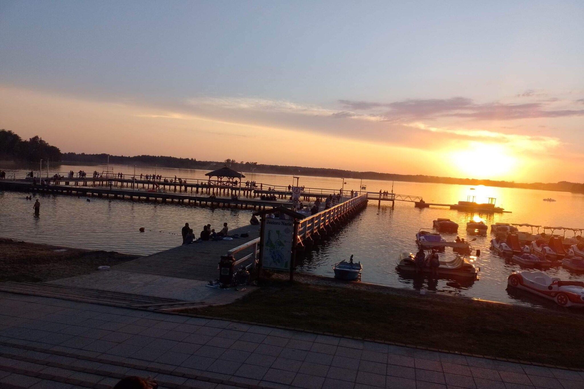 A waterfront pier with people and boats, a signboard, and a sunset.