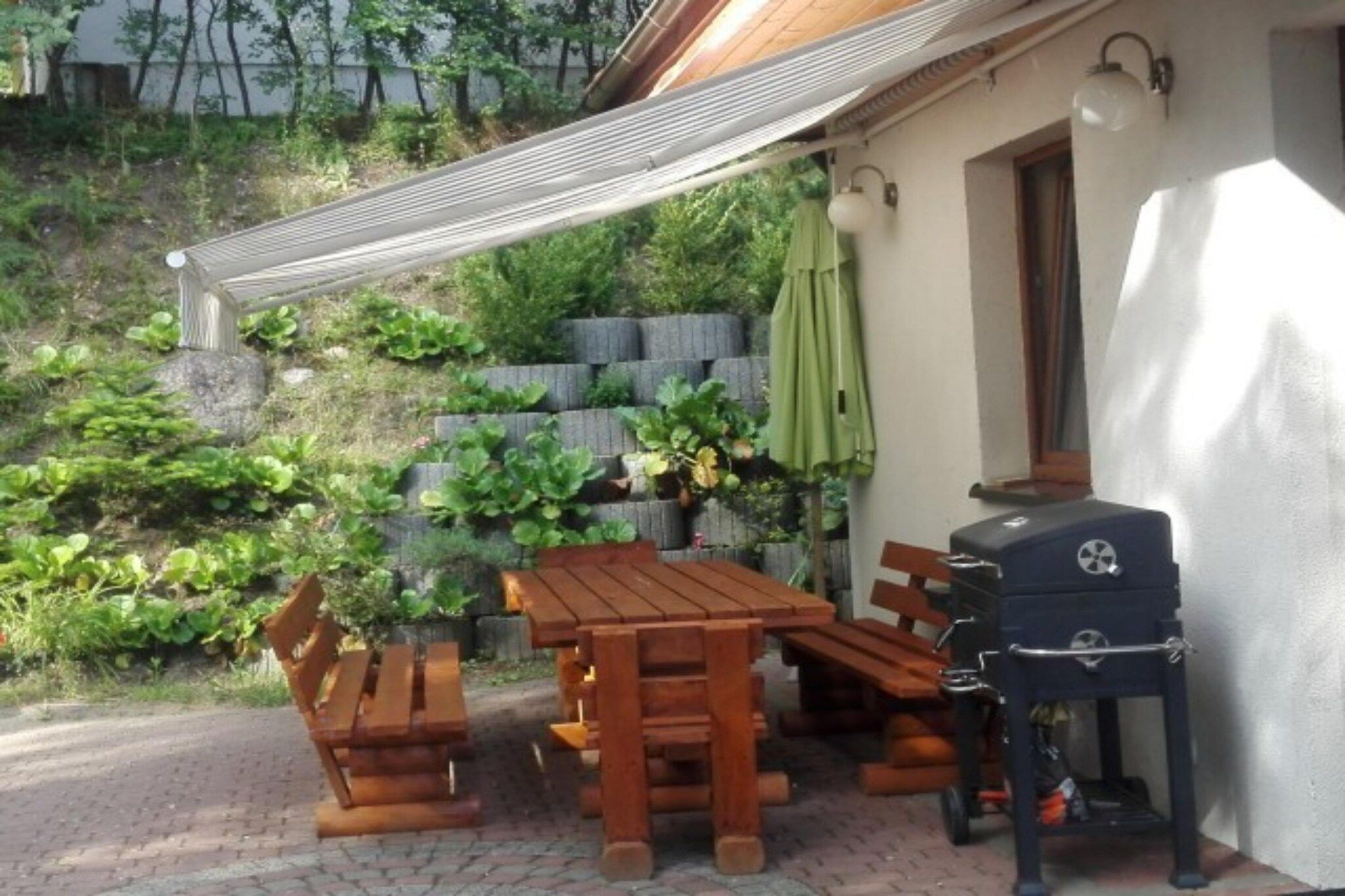 Een terras buiten met houten meubels, een barbecue en een stenen muur met beplanting.