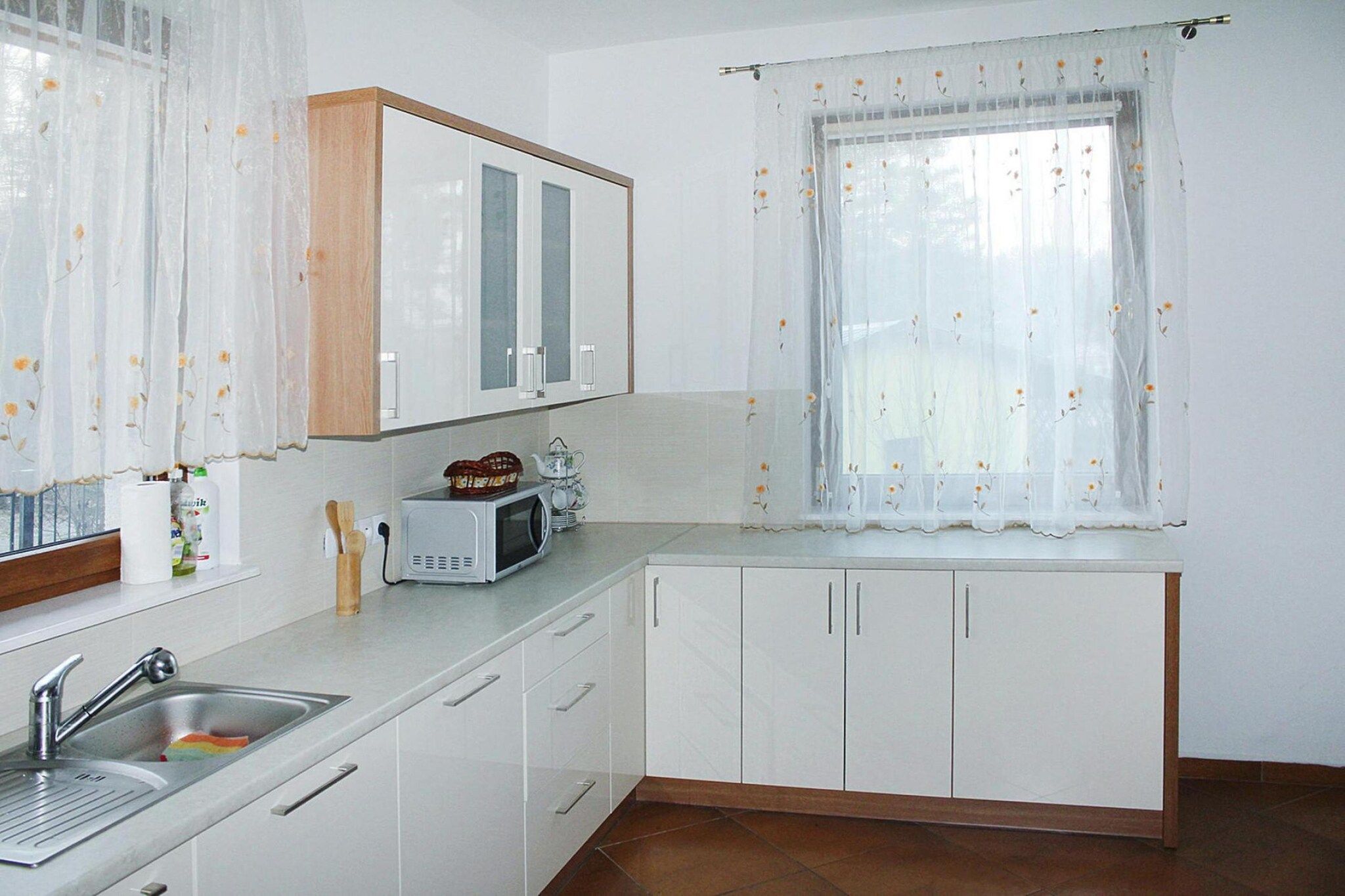 A kitchen with white cabinets, a microwave, a sink, and a window with floral curtains.