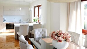 Una cocina moderna con armarios blancos, un comedor con mesa de vidrio y sillas blancas tapizadas, y un jarrón de flores sobre la mesa.
