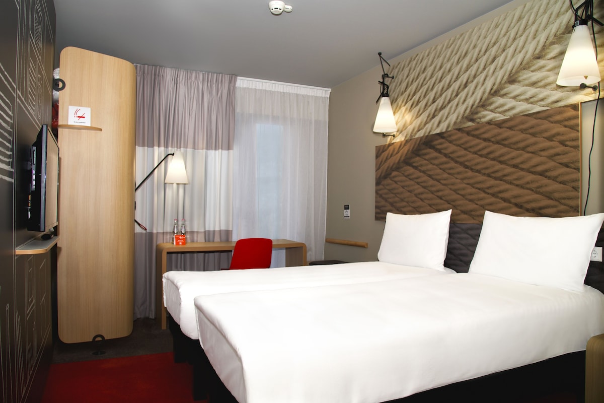 Una habitación de hotel con cama, escritorio, silla, televisor y lámpara.