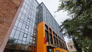 A modern building with a glass facade and orange accents, featuring a prominent entrance and outdoor seating area. - Ibis Tbilisi Stadium (Tbilisi)