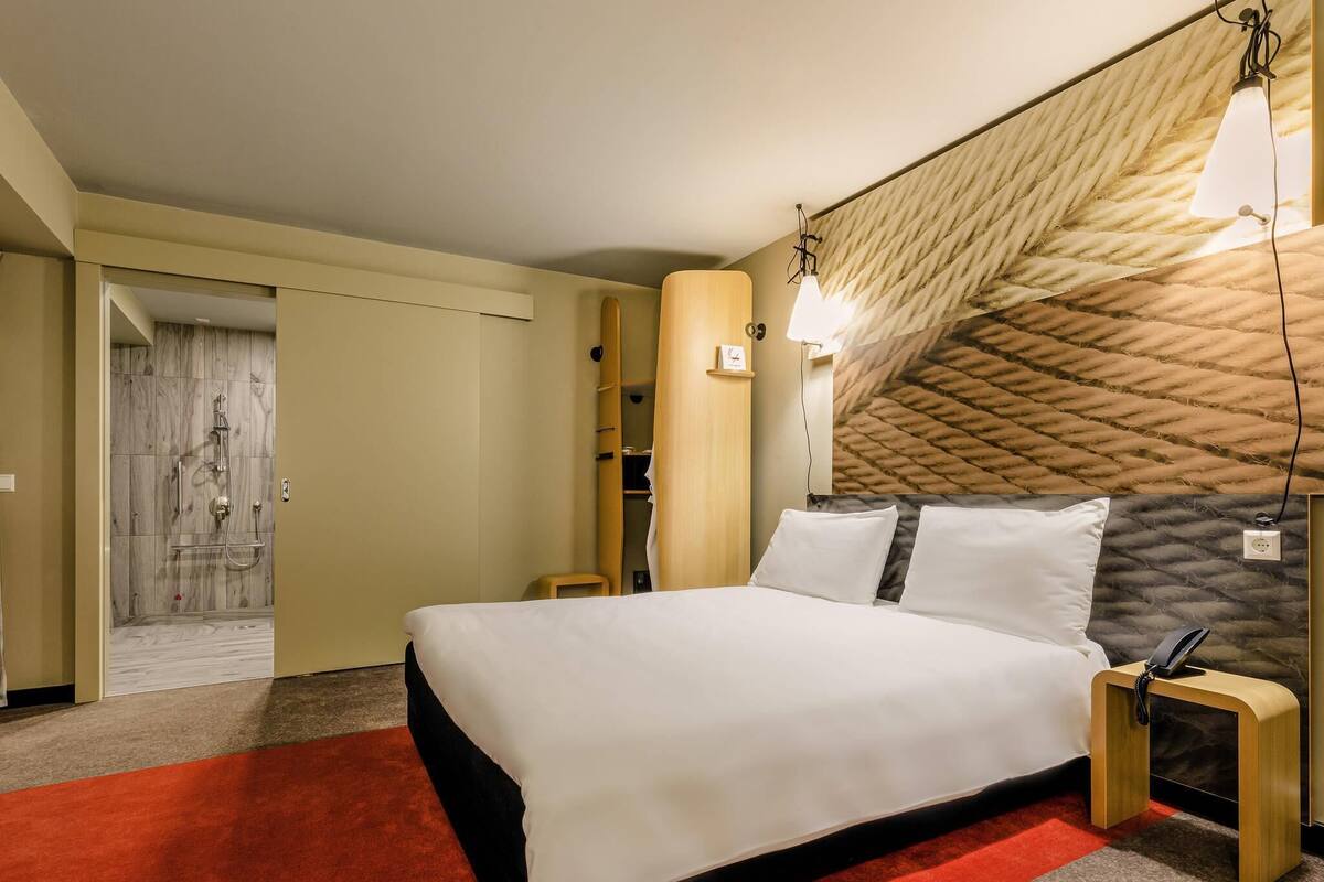 A hotel room with a bed, a red carpet, a wooden headboard, a shower area, and a wooden wardrobe.