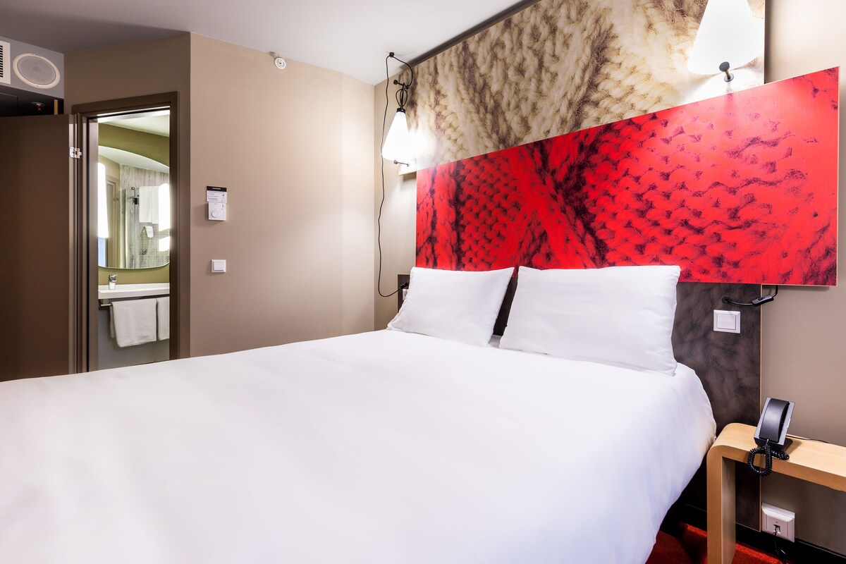 Una habitación de hotel con una cama grande, una mesita de noche con un teléfono y una pared con un estampado rojo.