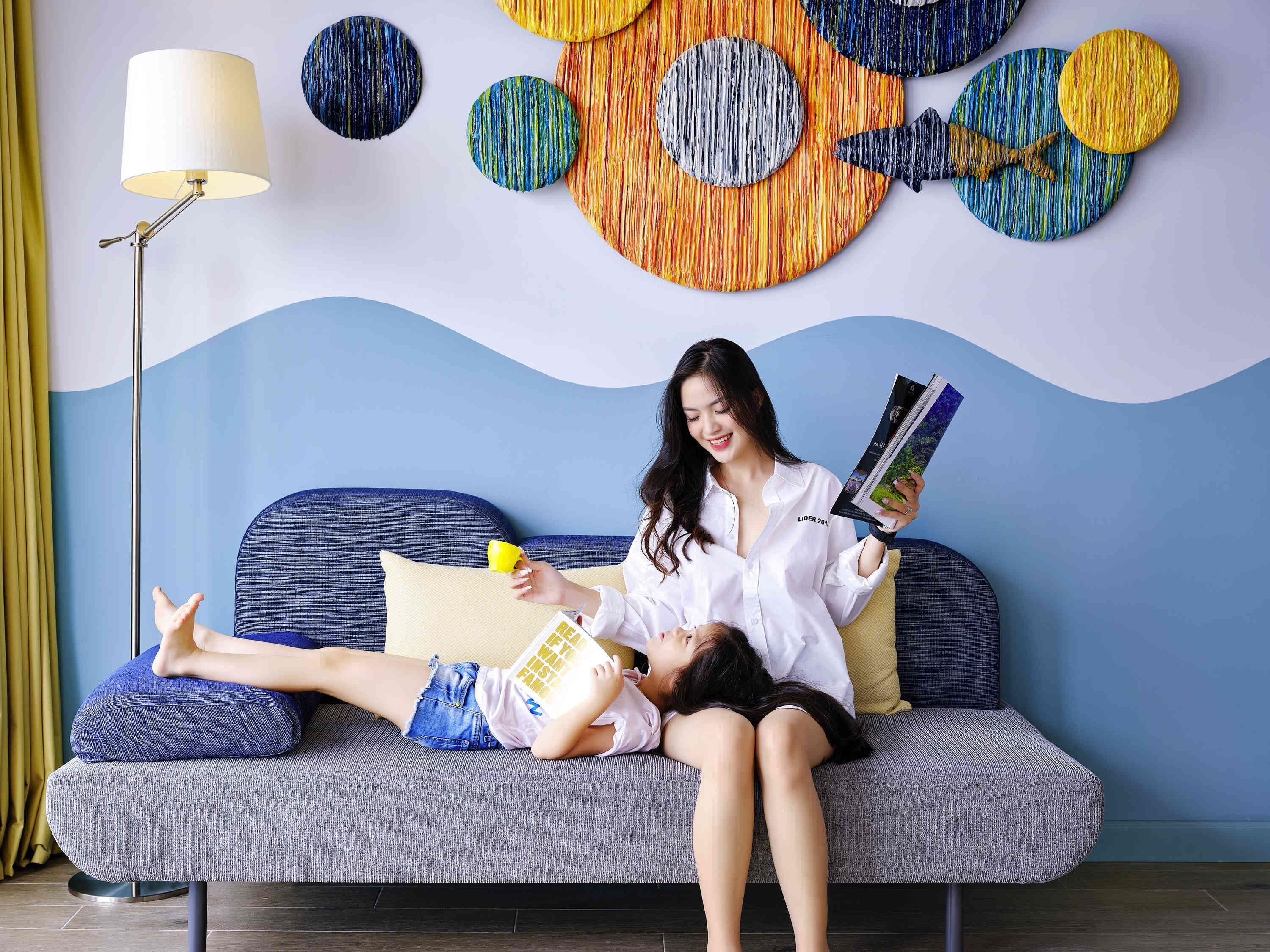 Une femme est assise sur un canapé, un enfant à ses côtés, et lit un magazine. On aperçoit également une lampe et des œuvres d’art abstraites accrochées au mur.