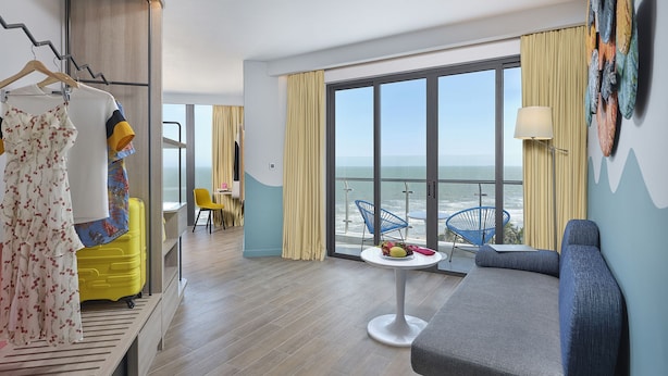 Una camera d'albergo moderna con un balcone, un'area salotto con un tavolo e un armadio con abiti appesi.