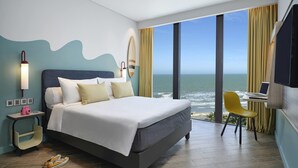 Una camera d'albergo moderna con un letto grande, una scrivania con un portatile, vista sull'oceano e una tavola da surf appesa al muro.