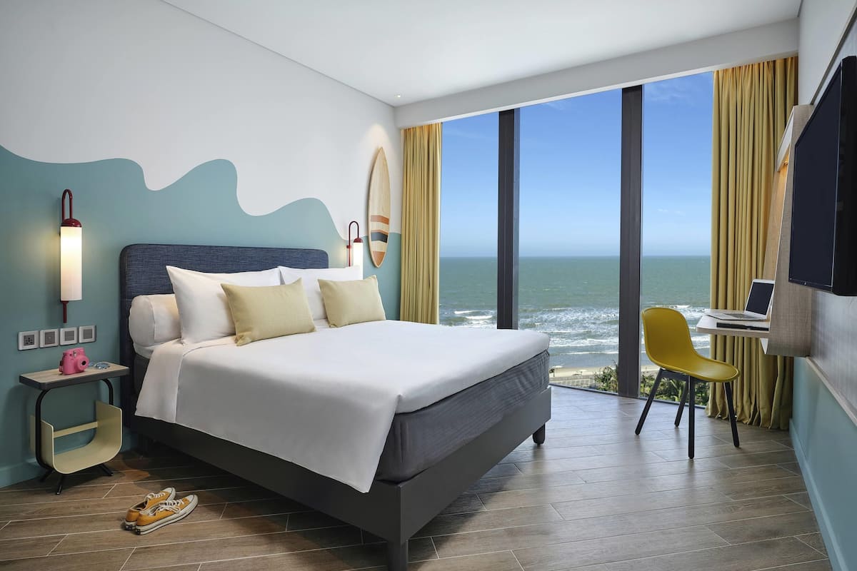 Ein modernes Hotelzimmer mit einem großen Bett, einem Schreibtisch mit einem Laptop, Blick auf den Ozean und einem Surfbrett an der Wand.