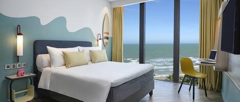 A modern hotel room with a large bed, a desk with a laptop, a view of the ocean, and a surfboard on the wall.