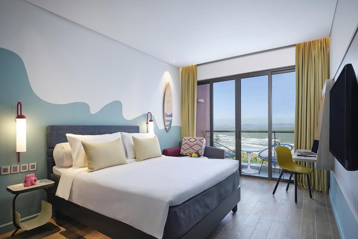 Ein modernes Hotelzimmer mit einem großen Bett, einem Schreibtisch mit einem Laptop, einem Sessel und einem Balkon mit Blick.