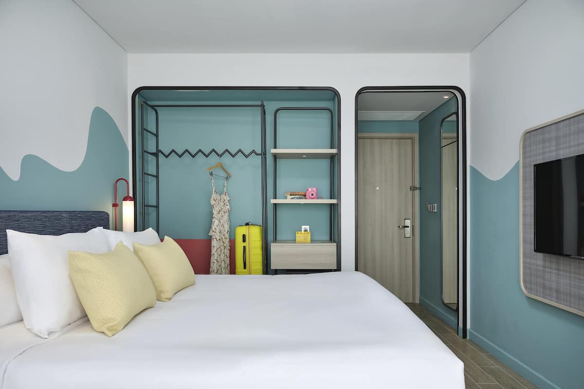 Ein modernes Hotelzimmer mit einem großen Bett, einem Kleiderschrank mit Spiegel, einem Ablagefach für Toilettenartikel und einem an der Wand befestigten Fernseher.