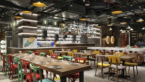 Interno di un ristorante con diversi tavoli, sedie colorate e un’area bar con utensili da cucina esposti alla parete.
