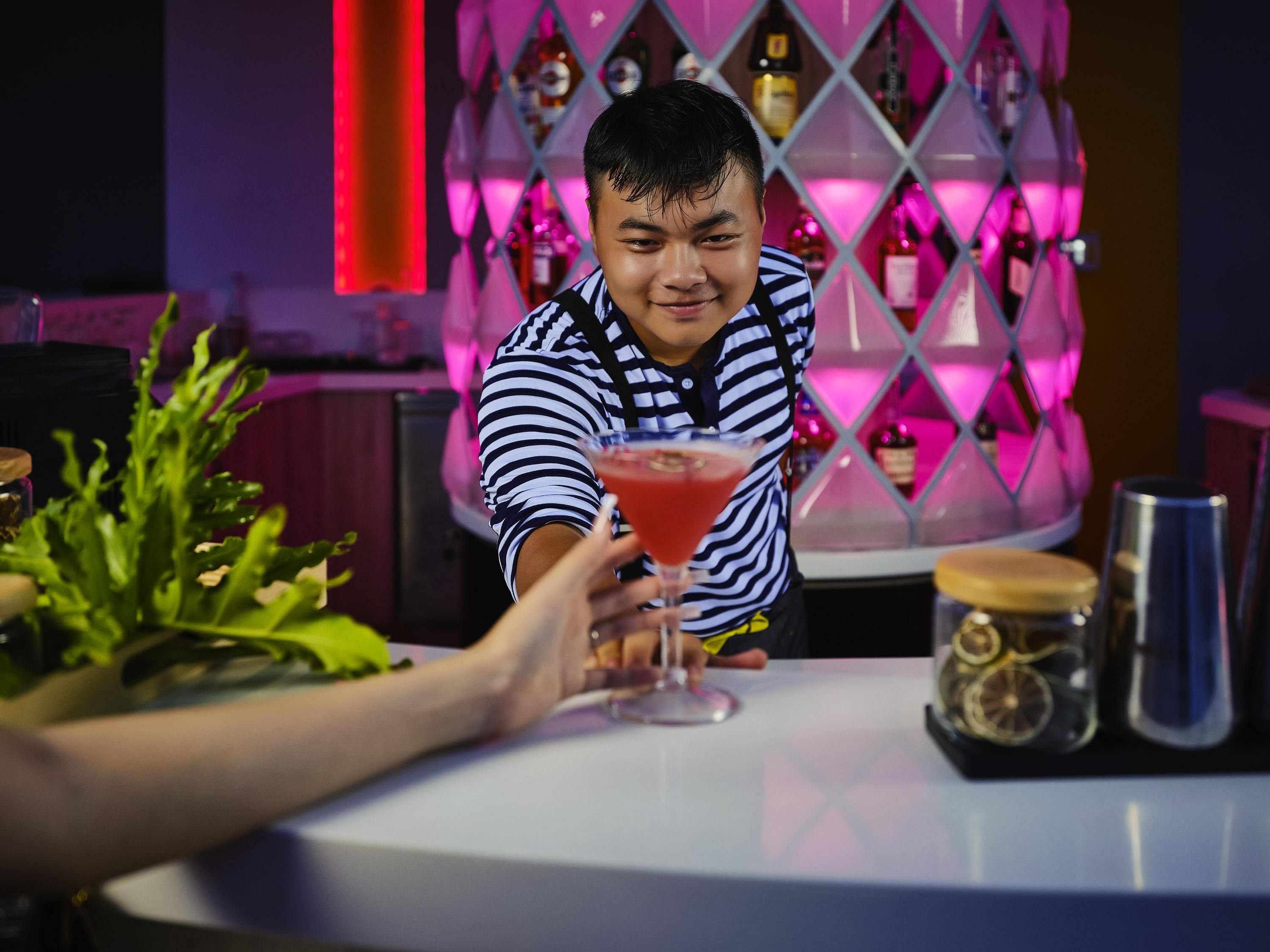 A person holding a red cocktail glass at a bar with a diamond-patterned backdrop and various bottles.