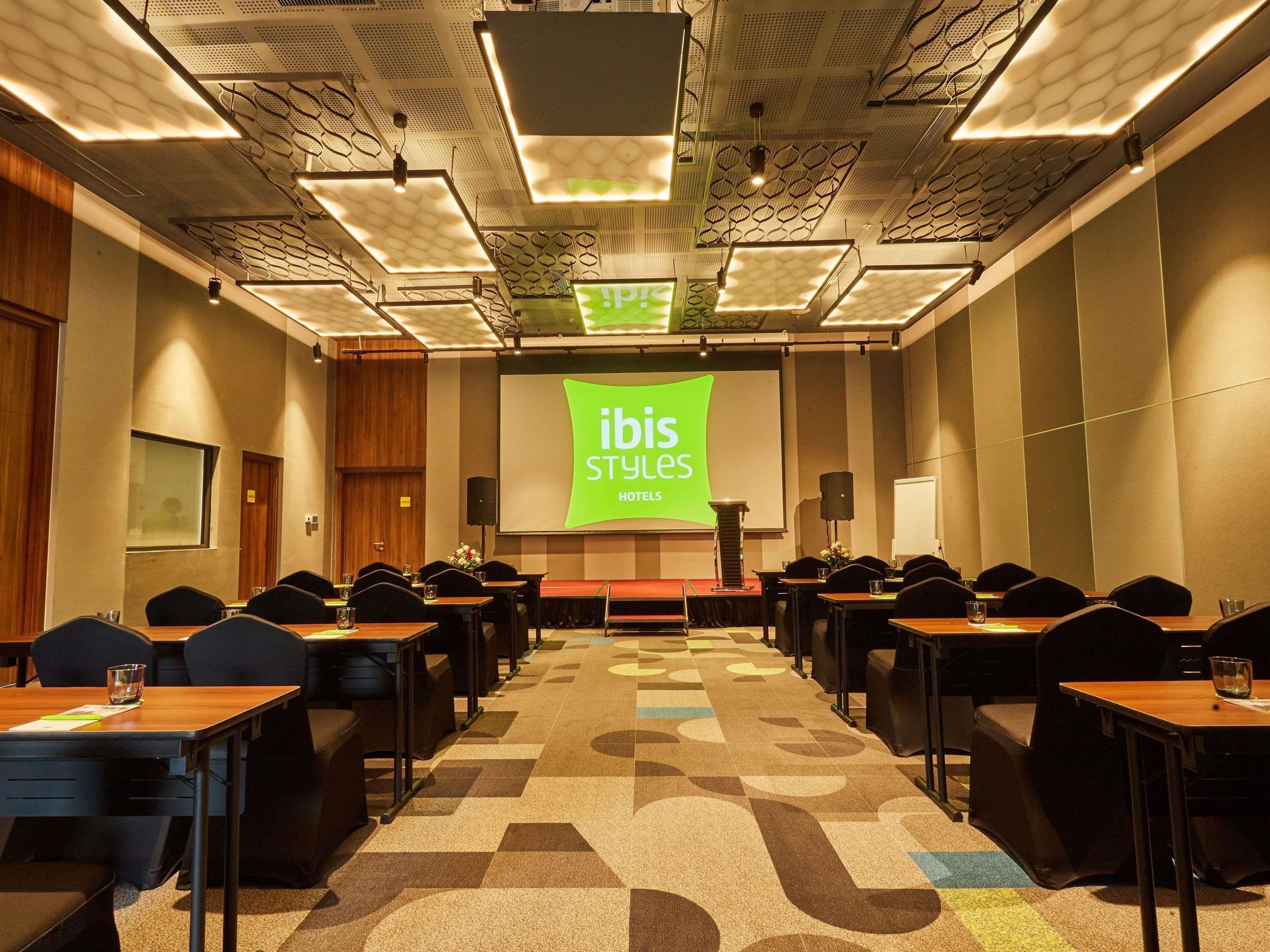 A conference room with a stage, multiple tables, and chairs, and a projection screen displaying 'ibis Styles Hotels'.