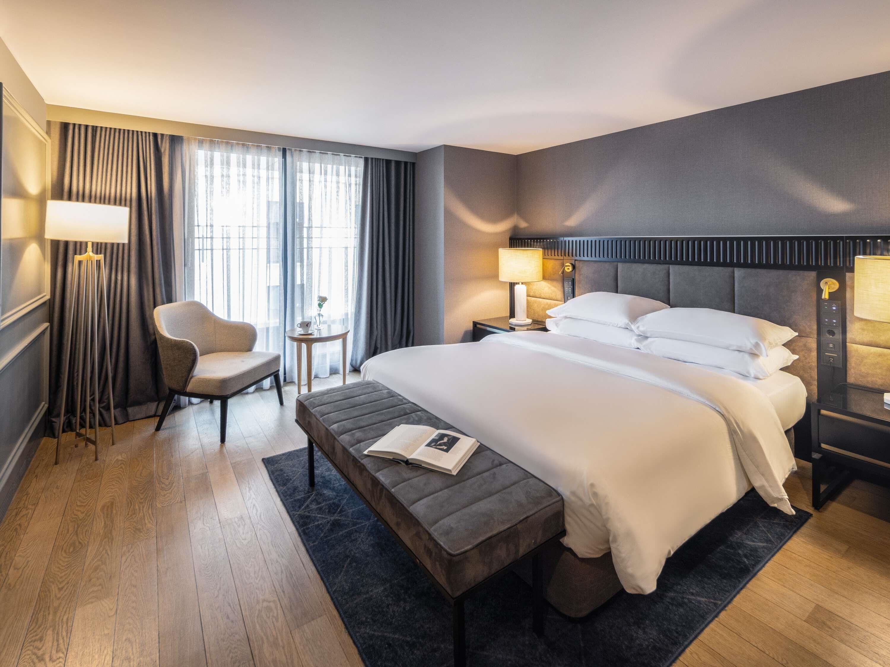 A hotel room with a bed, bedside tables, a chair, a small table with a lamp, and a window with curtains.
