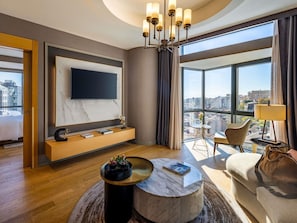 Moderni hotellihuone, jossa on suuri televisio, pyöreä sohvapöytä, sohva ja näkymä kaupunkiin.