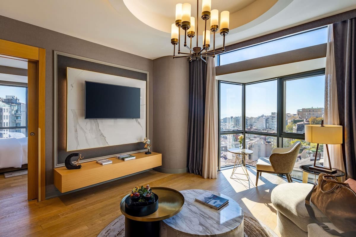Habitación de hotel moderna con una televisión grande, una mesa de centro redonda, un sofá y vistas a la ciudad.