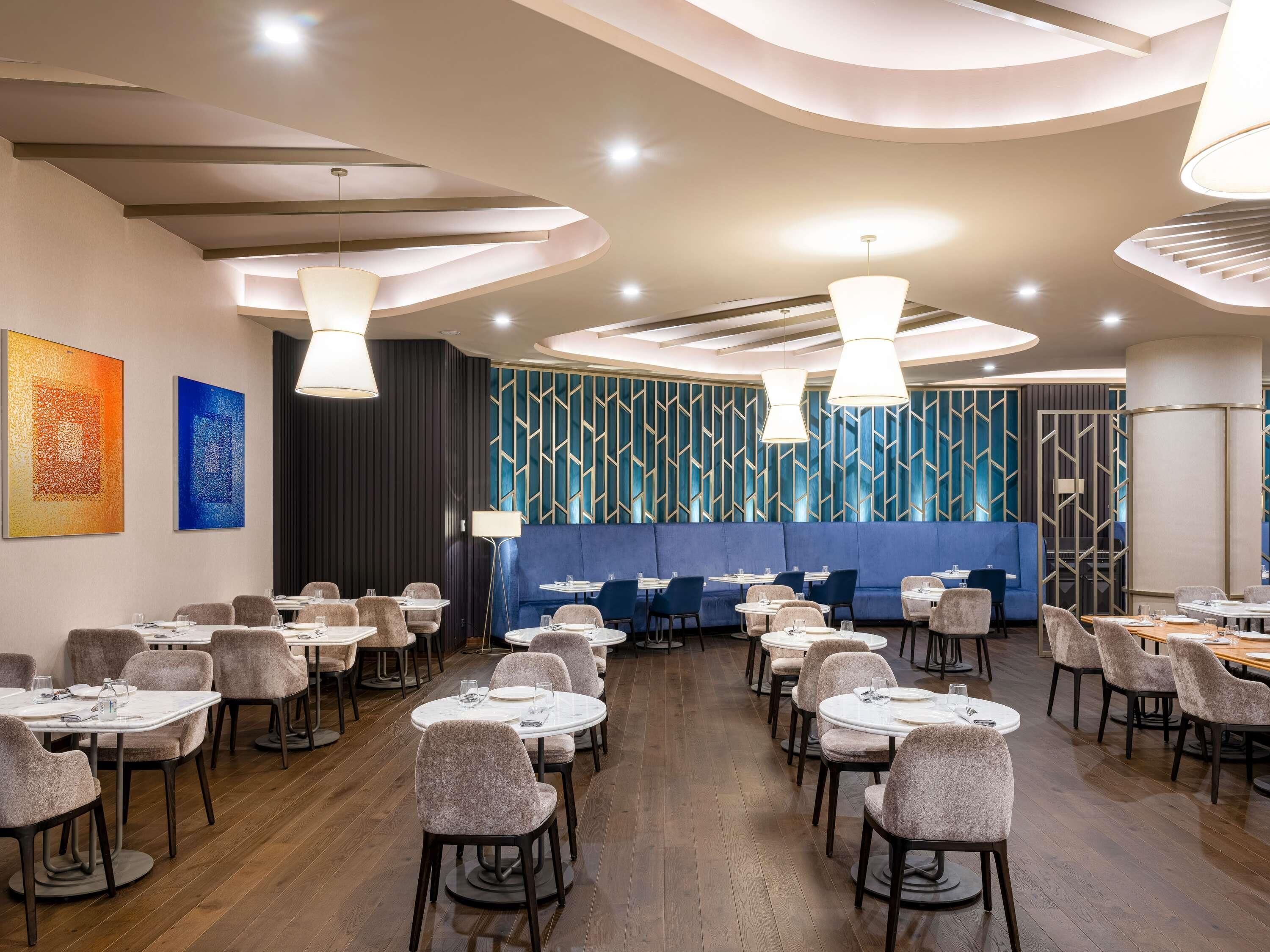Un comedor moderno con mesas y sillas redondas, una zona de asientos tipo banca en azul y arte abstracto en las paredes.