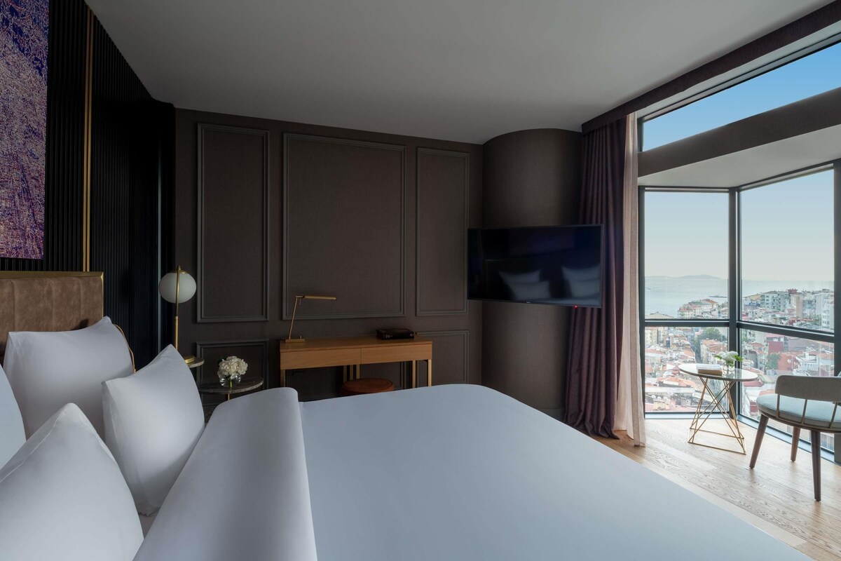 A modern hotel room with a large bed, a desk, a flat-screen TV, and a city view.