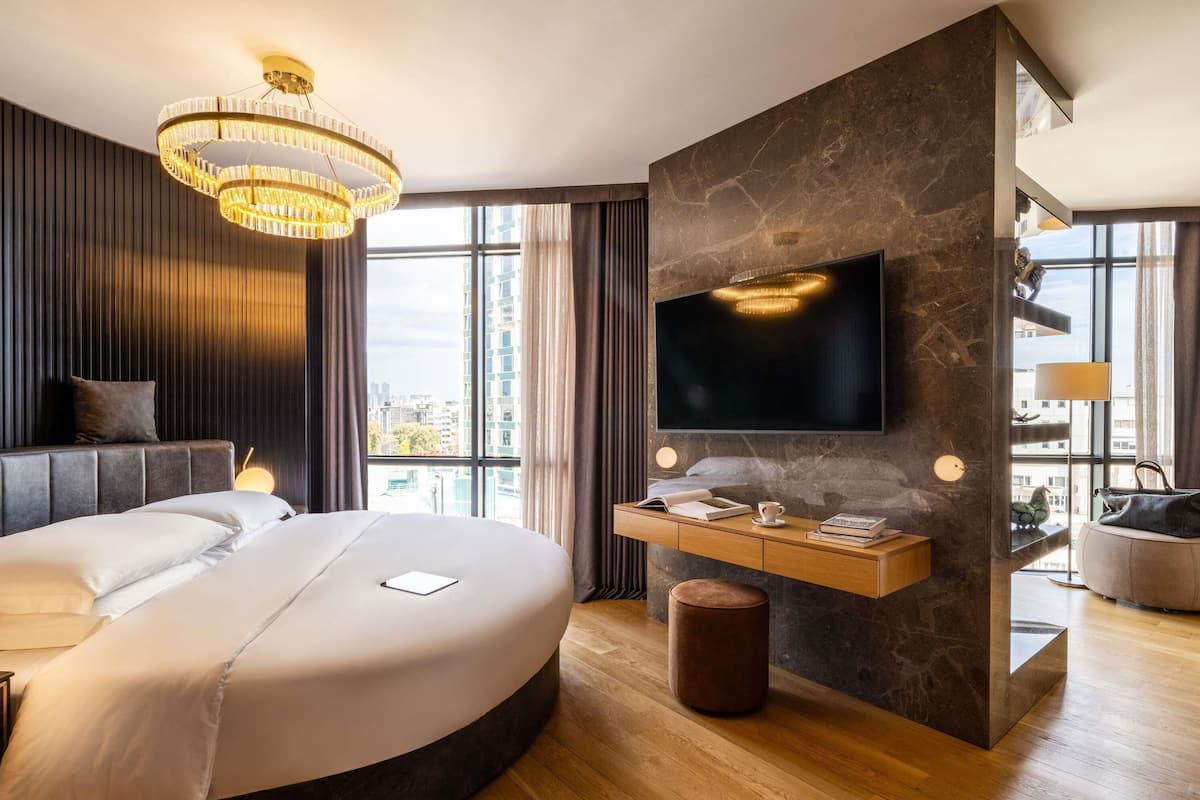A modern bedroom with a large bed, a TV mounted on a stone wall, and a city view.