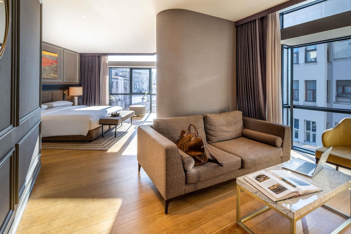 A hotel room with a bed, a sofa, a coffee table with a laptop and book, and a large window with a city view.