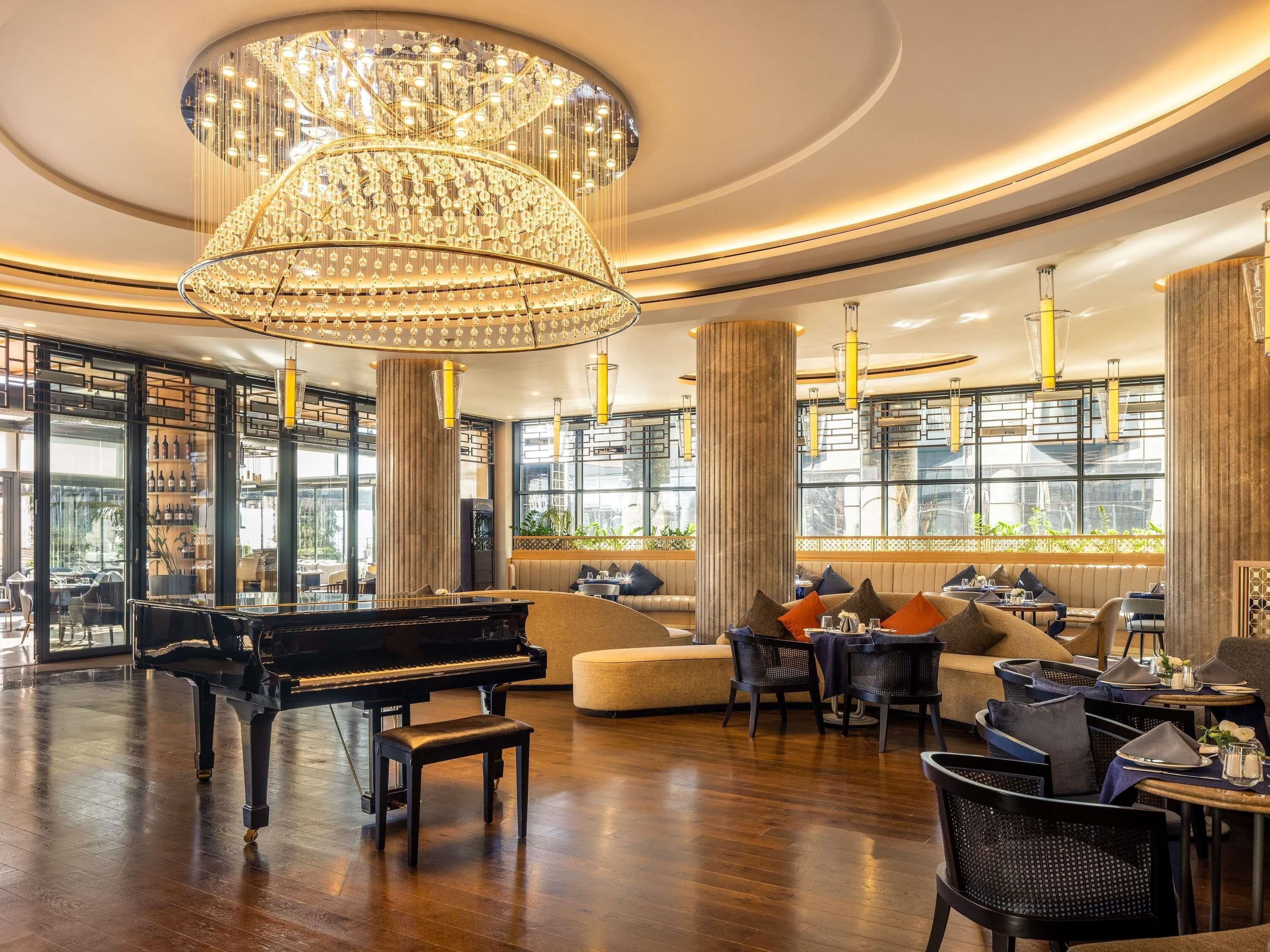 Un magnifique piano à queue trône dans un salon spacieux et élégamment aménagé, agrémenté d’un grand lustre, de fauteuils confortables et de larges fenêtres.