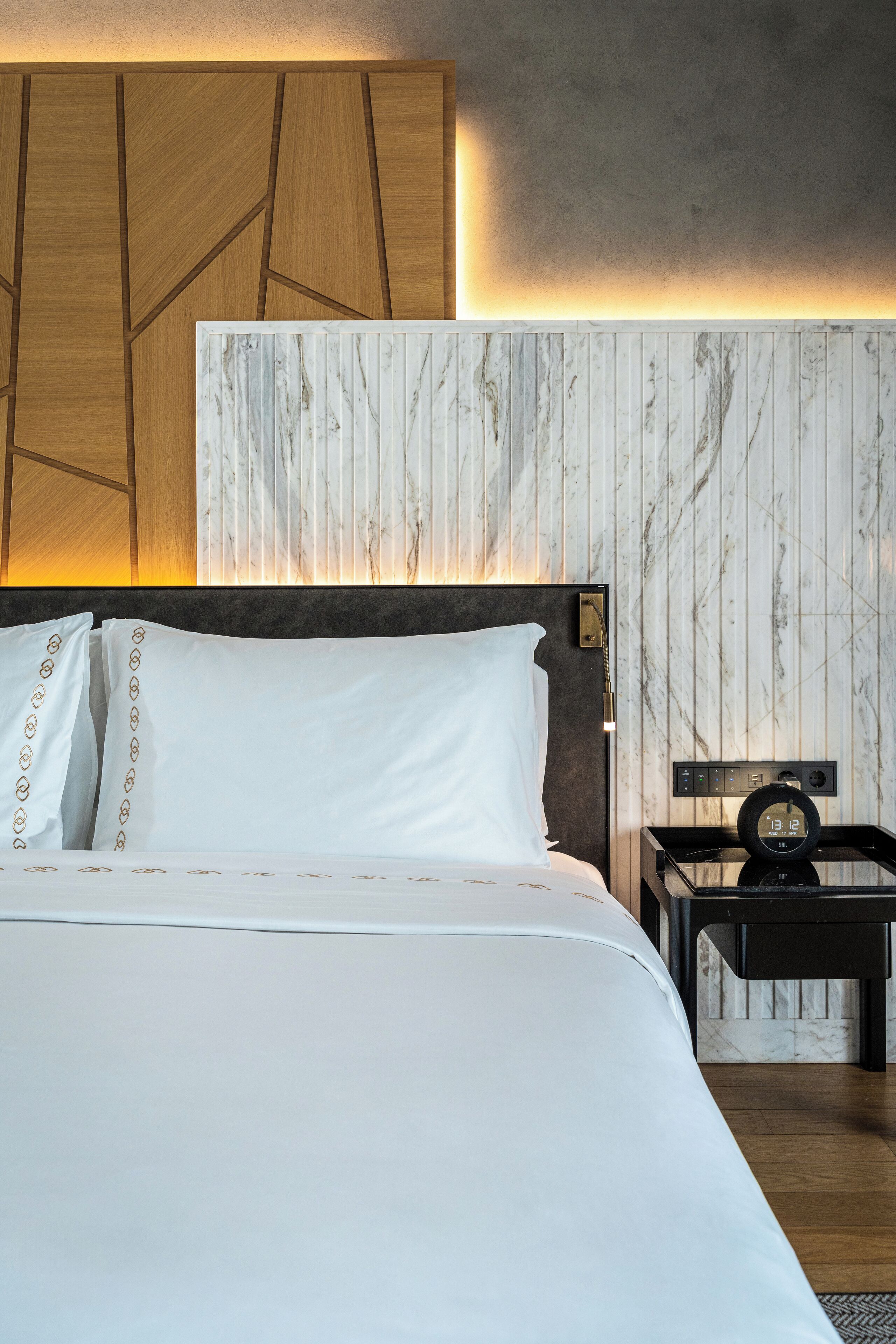 A neatly made bed with white linens and decorative pillows, a bedside table with a clock, and a wall with a geometric wooden panel and a marble-like textured panel.