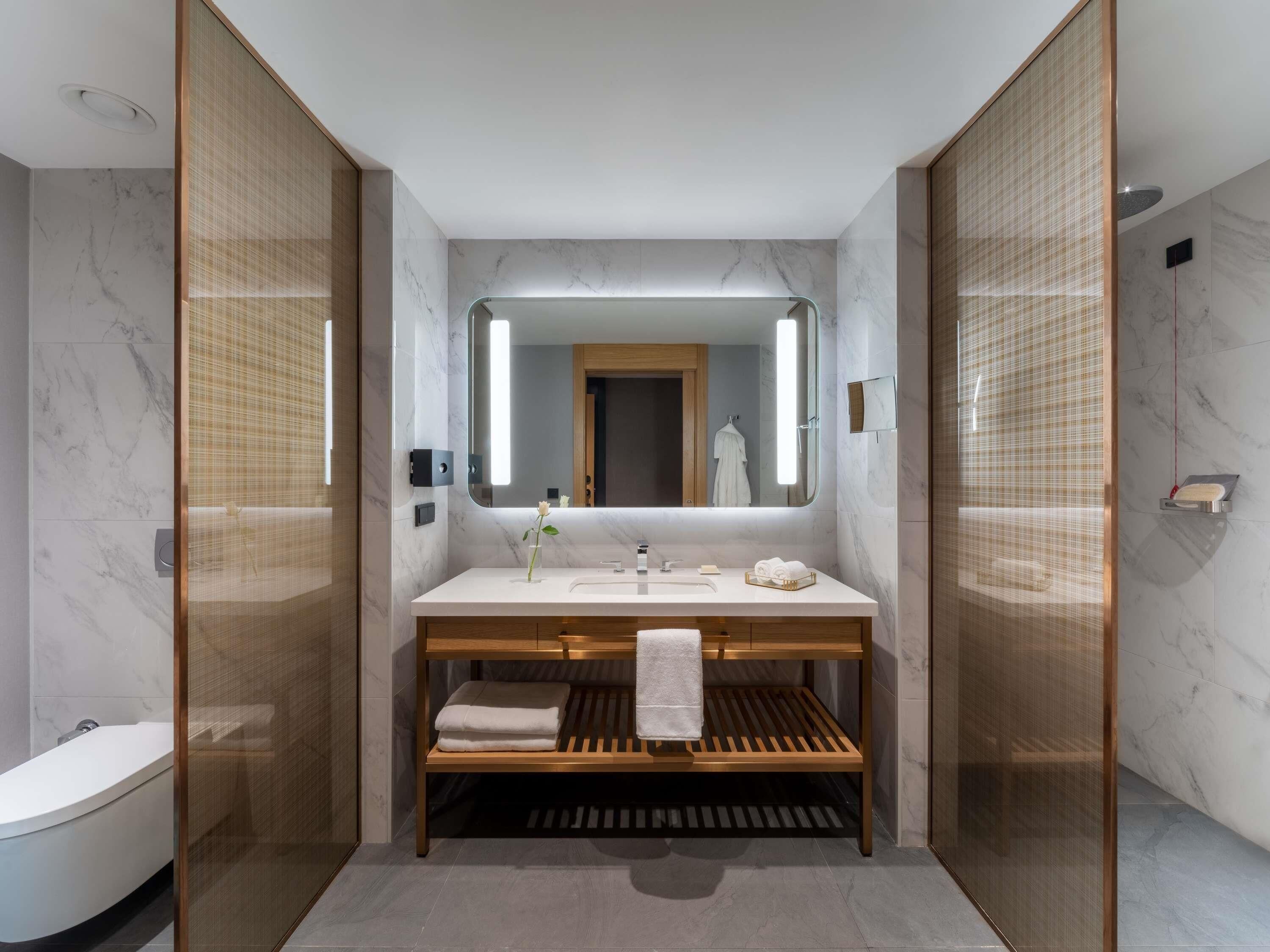 A modern bathroom with a marble vanity, a large mirror, and a wooden bench.