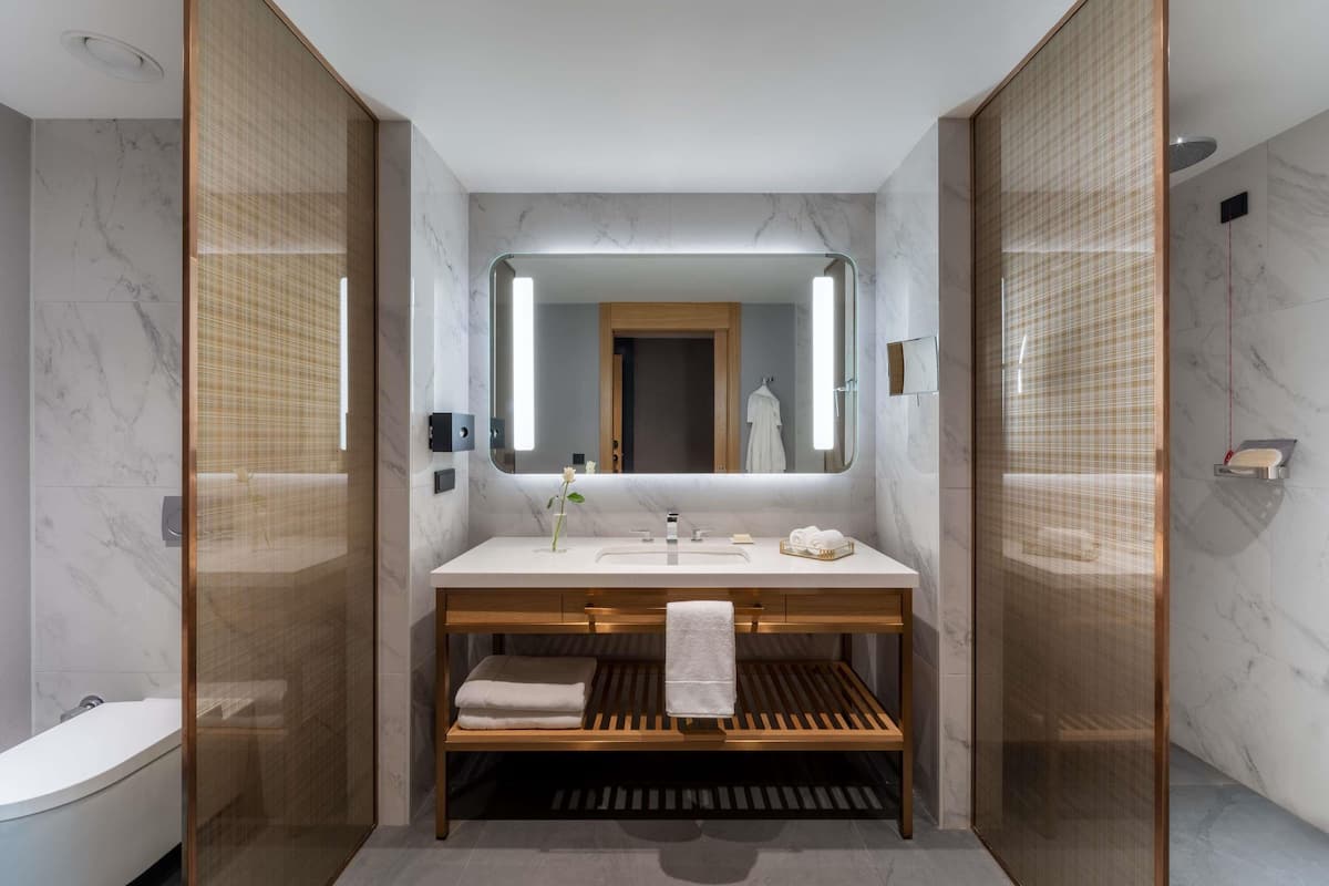 A modern bathroom with a marble vanity, a large mirror, and a wooden bench.