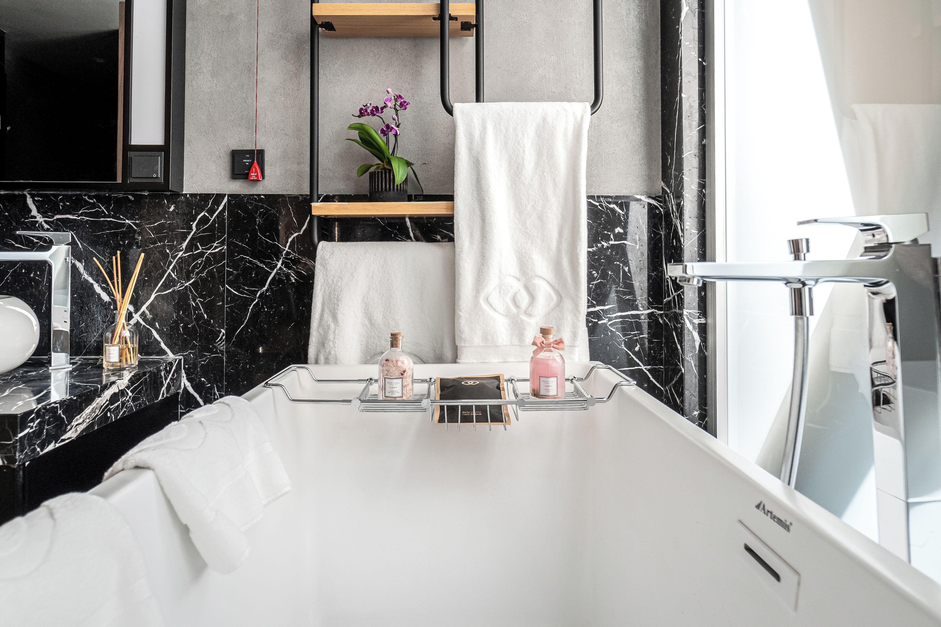 Een moderne badkamer met een witte badkuip, zwarte marmeren tegels, een witte handdoek en twee flessen zeep op een plank.