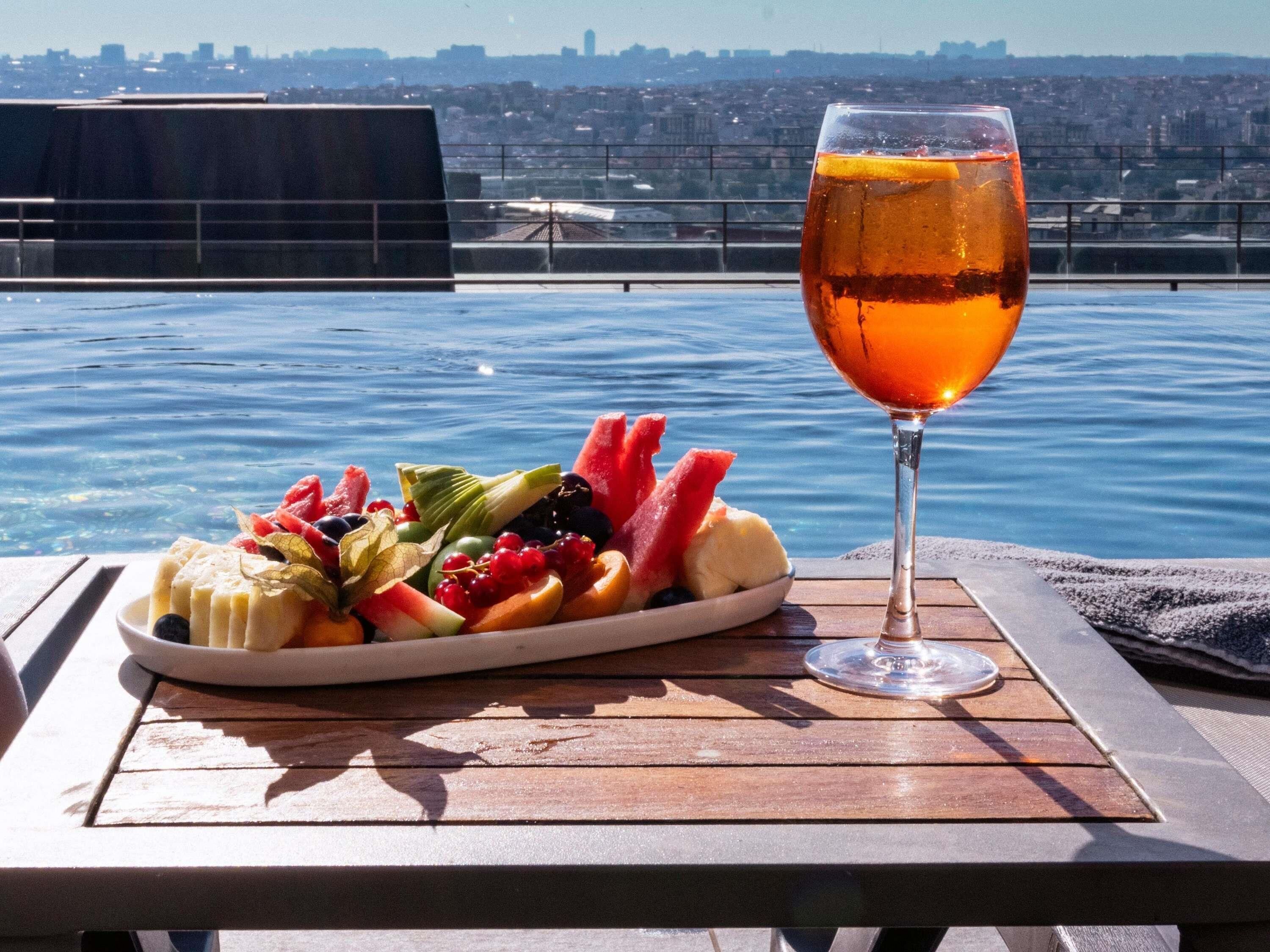 Un tavolo con un vassoio di frutta e un bicchiere di bevanda ambrata, con vista su un corpo d'acqua e sullo sfondo un panorama urbano.