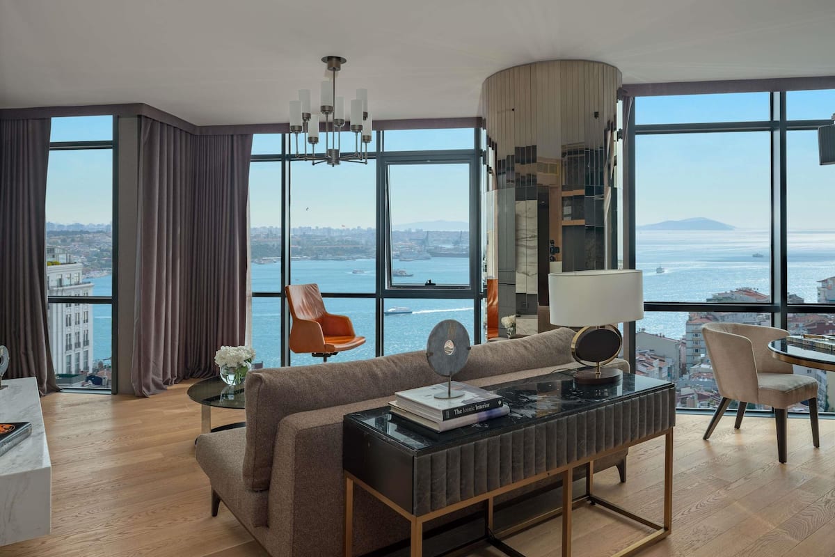 Una sala de estar moderna con sofá, mesa de centro y sillones, ventanas amplias con vista a la ciudad y al agua, y una araña.