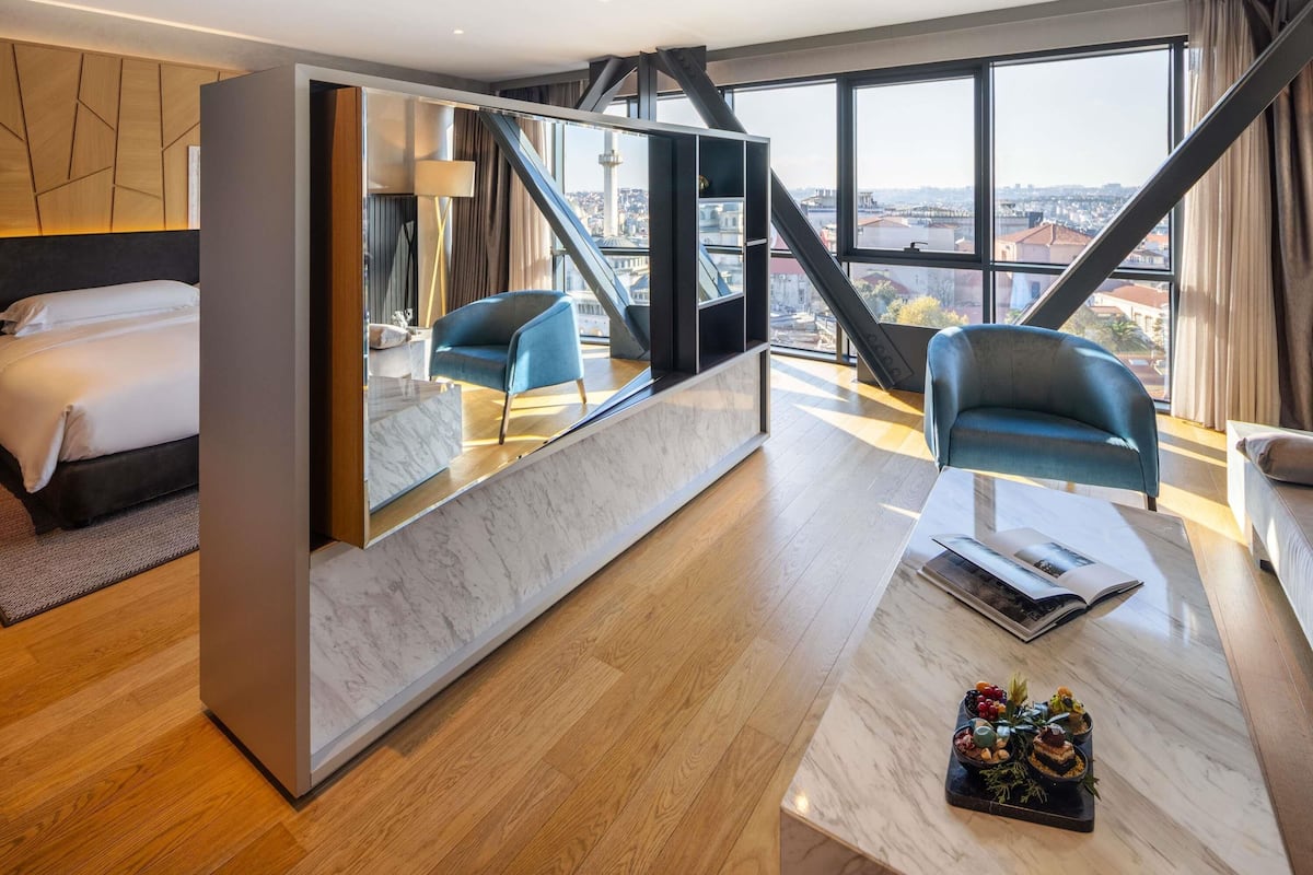 Una habitación de hotel moderna con una cama, un área de descanso con una mesa y un gran ventanal con vistas a la ciudad.