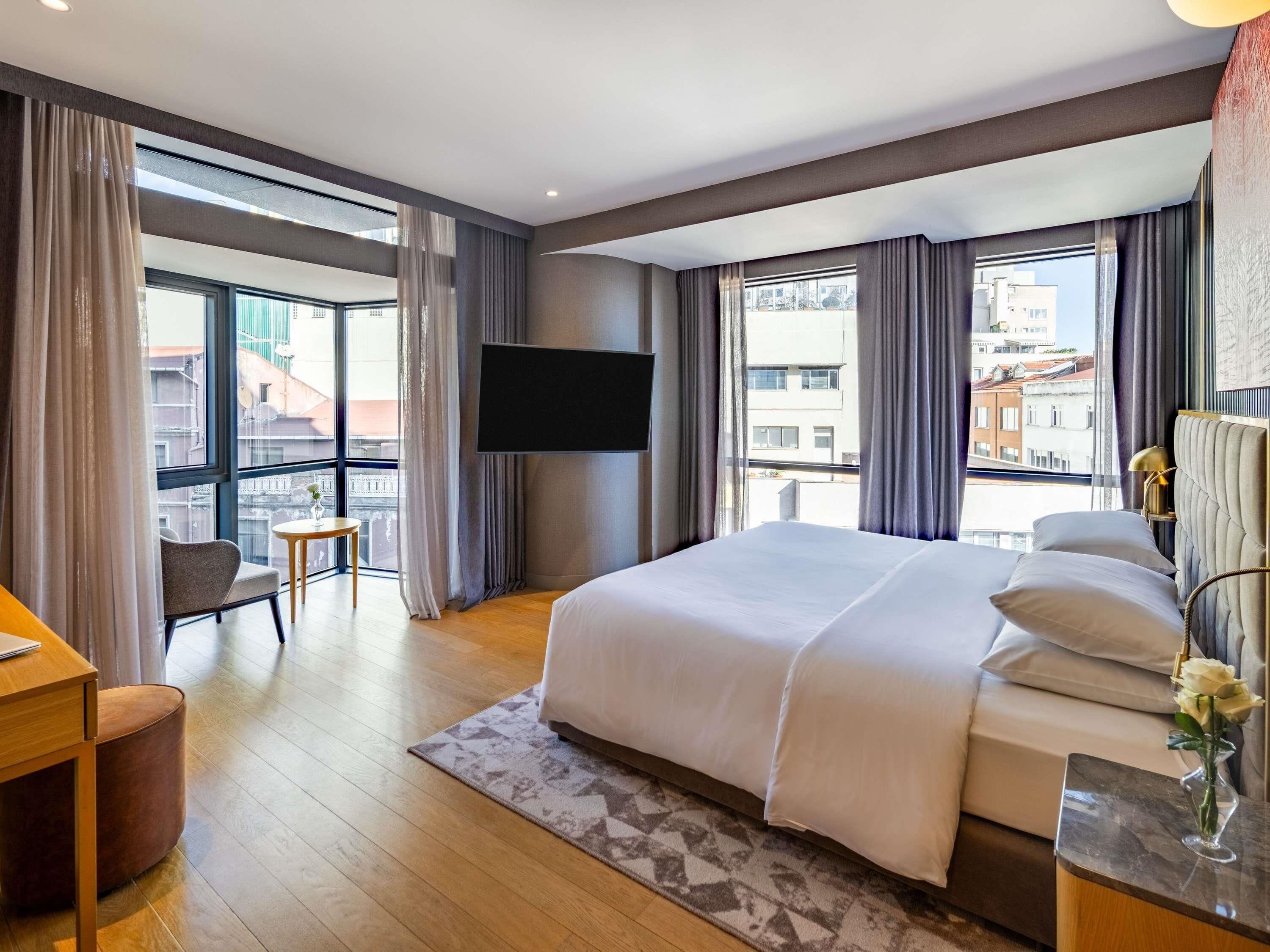 A modern hotel room with a large bed, a TV, a desk, and a view of the city.