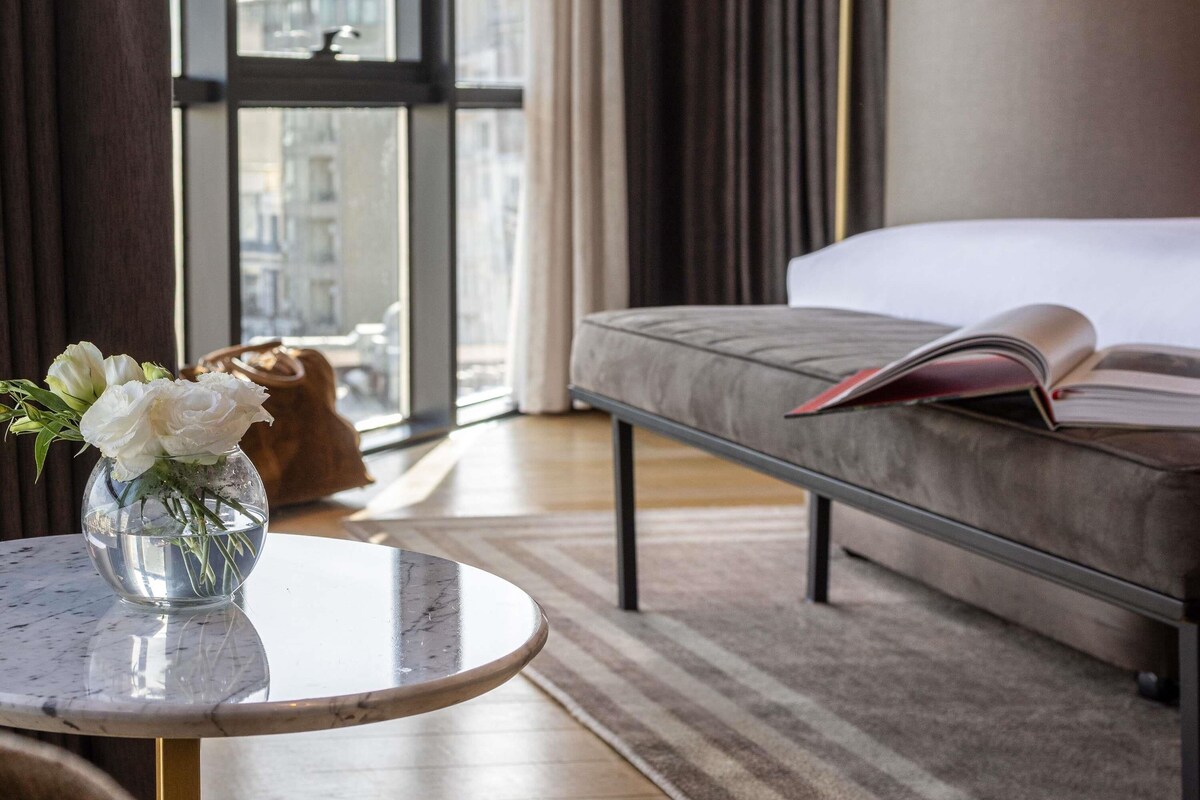 A hotel room with a bed, a bedside table with a vase of flowers, a window with curtains, and a view of buildings outside.