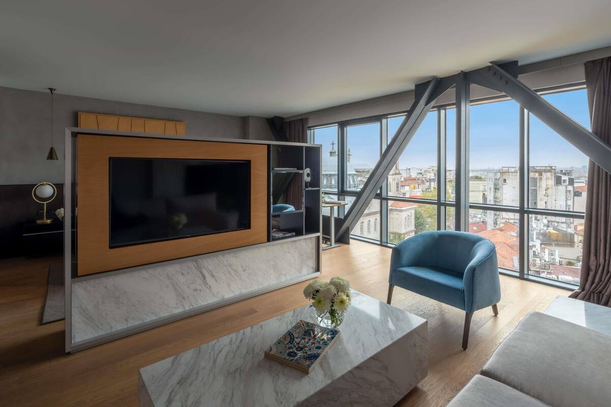 Een moderne woonkamer met een grote televisie, een blauwe fauteuil, een marmeren salontafel en uitzicht op de stad door grote ramen.