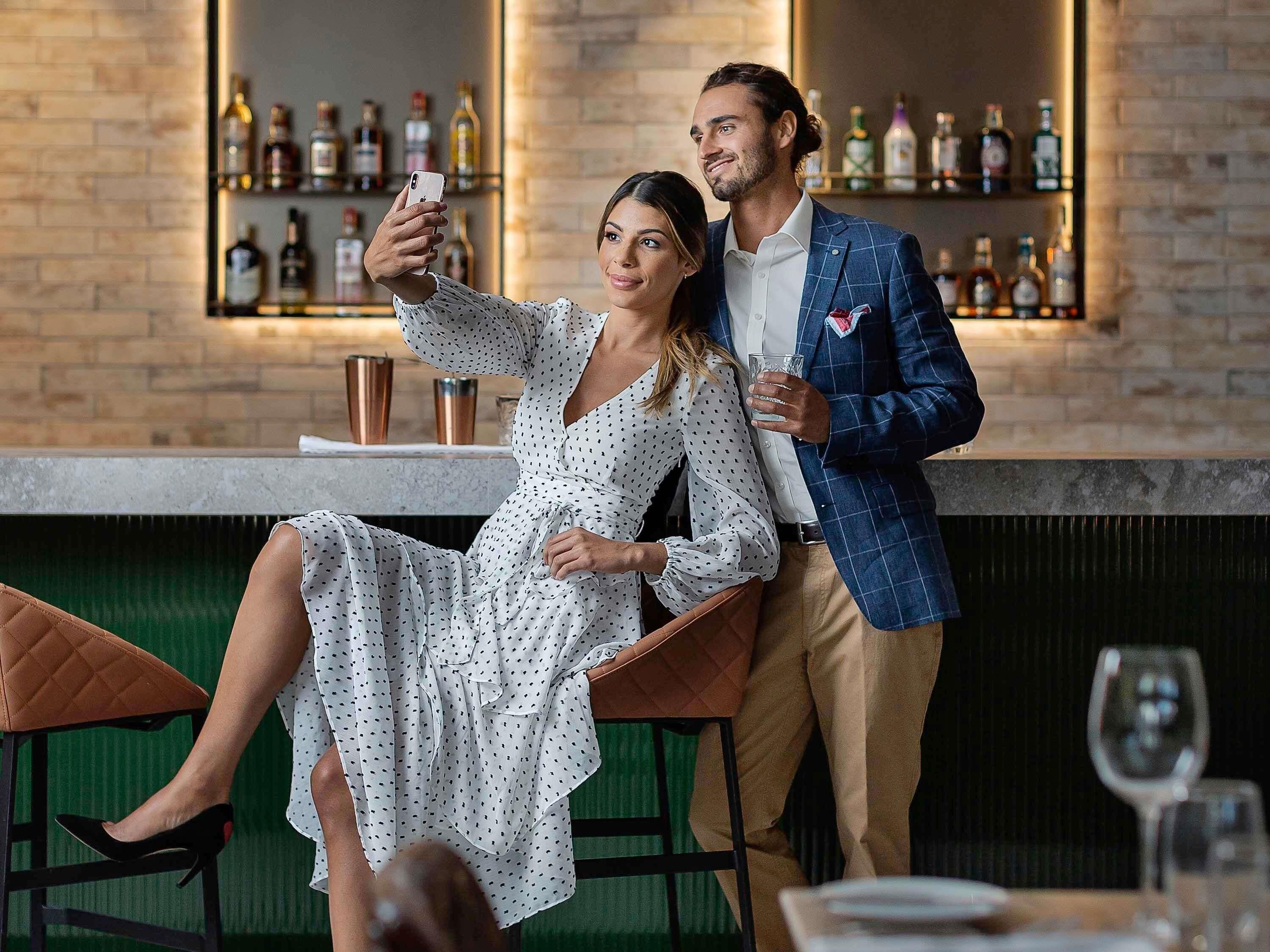 Un coin bar avec un mur en briques, des étagères remplies de bouteilles, et un couple qui prend un selfie.