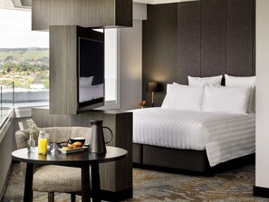 A modern hotel room with a bed, a TV mounted on the wall, a small table with breakfast items, and a view of the countryside.