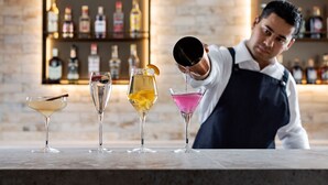 A bartender pouring a drink into a martini glass at a bar with various bottles in the background.