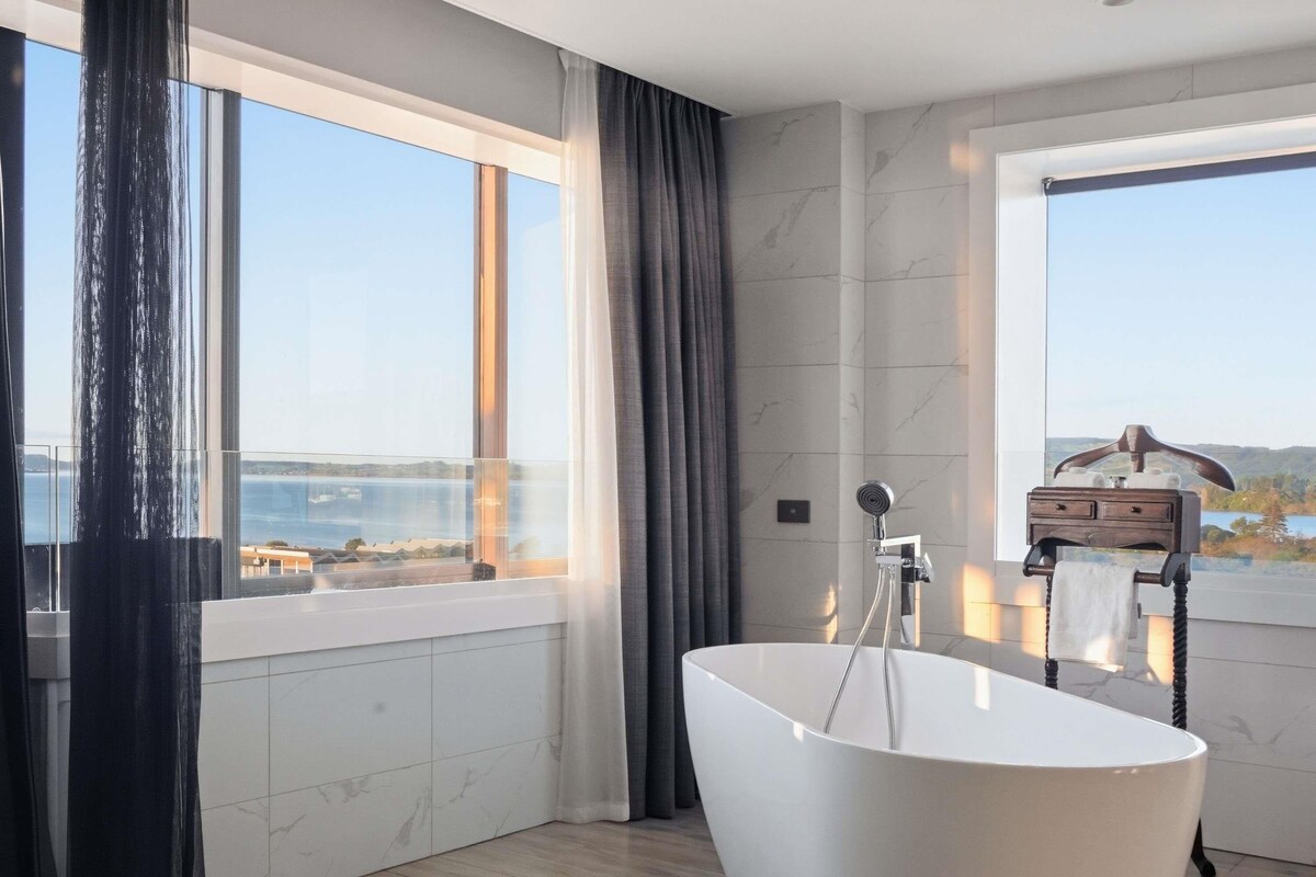 A modern bathroom with a freestanding bathtub, large windows, and a view of the sea.