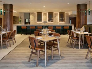 A modern restaurant with wooden tables and chairs, a bar area with shelves of bottles, and a stone fireplace.