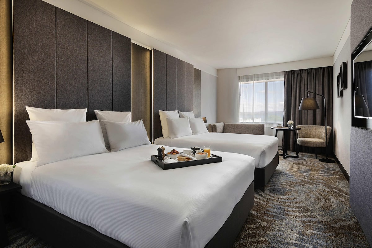 Habitación de hotel con dos camas, un área de descanso, una pequeña mesa con provisiones para el desayuno y un amplio ventanal con cortinas.