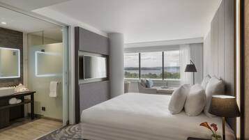 A modern hotel room with a large bed, a TV, a bathroom with a glass shower, and a view of the city.