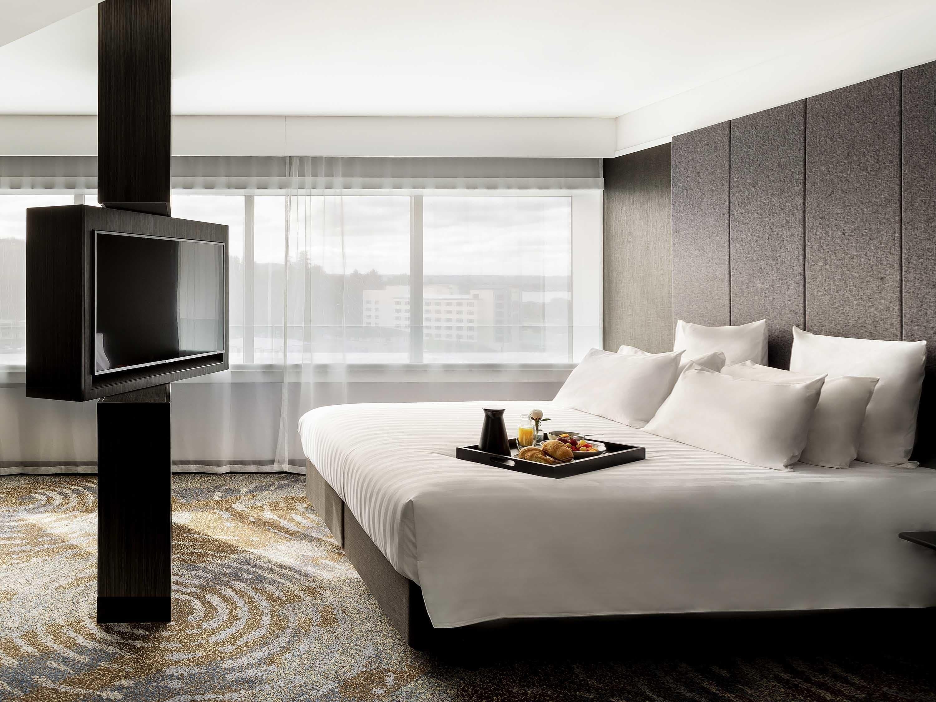 A hotel room with a bed, a flat-screen TV mounted on a dark pillar, a breakfast tray on the bed, and a large window with a city view.