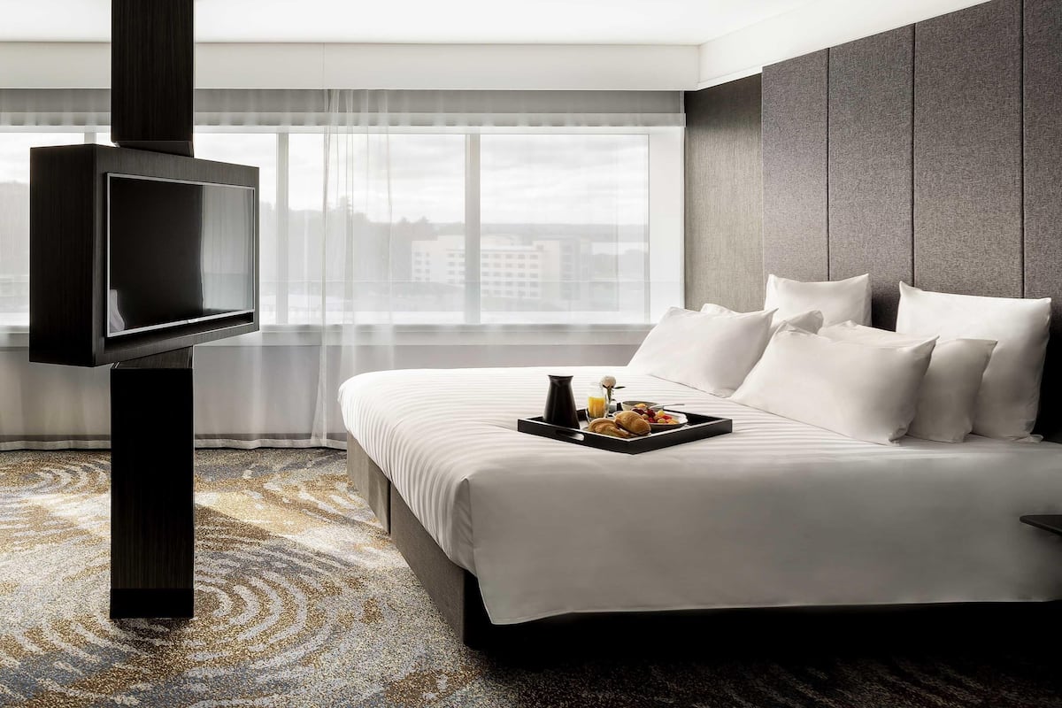 A hotel room with a bed, a flat-screen TV mounted on a dark pillar, a breakfast tray on the bed, and a large window with a city view.