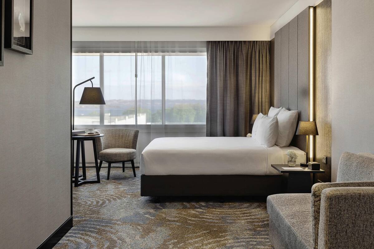 Habitación de hotel con cama, dos sillas, una mesita, una lámpara y vistas al exterior.