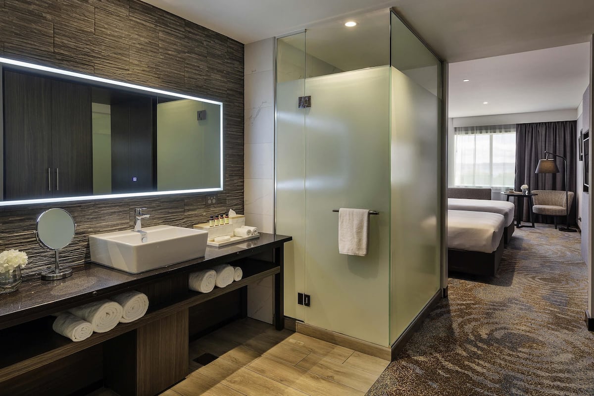 A modern hotel bathroom with a large mirror, a sink, a glass-enclosed shower, and a bed in the adjacent room.