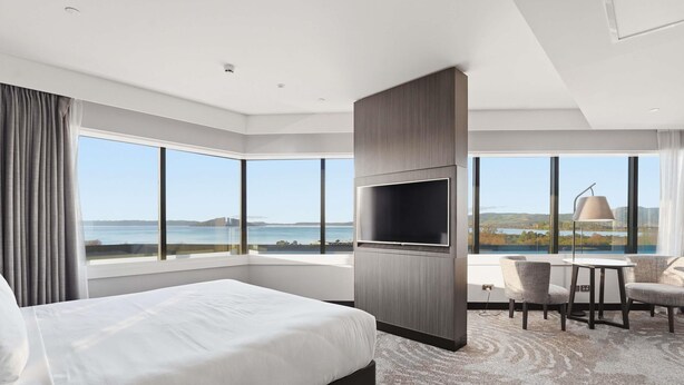 A modern hotel room with a large bed, a flat-screen TV, a dining area with chairs, and a view of the sea.